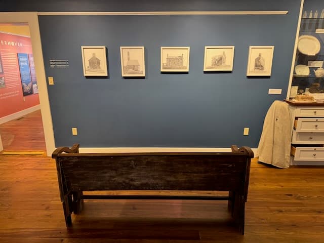 An old dark wooden church pew displayed against a blue wall hung with five framed photographs of former slave dwellings.