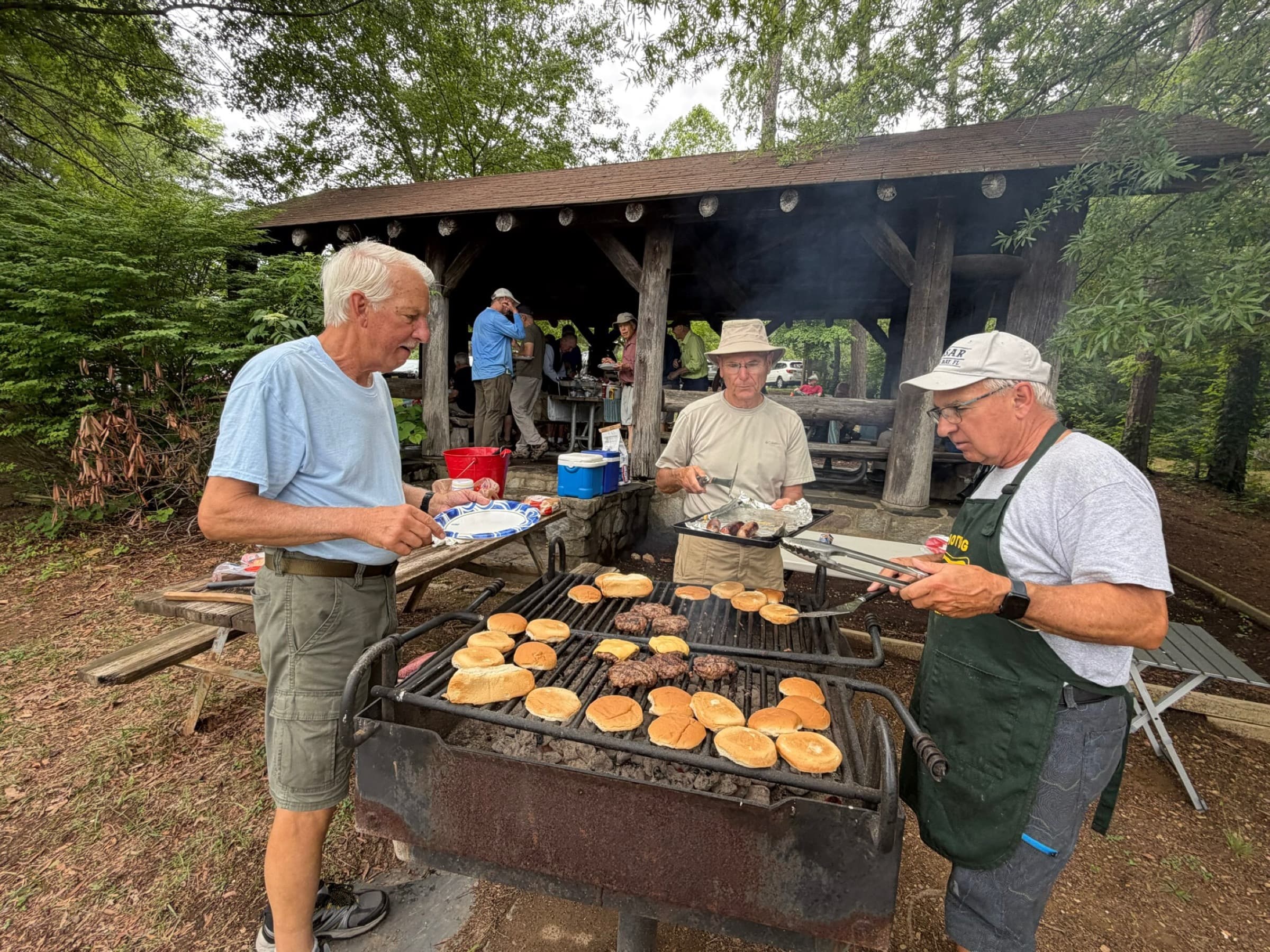 Three hikers grilling hamburgers and buns on a charcoal grill beside the park shelter.